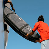 Roam Surfboard Converter Coffin grau-schwarz, zwei Personen laden Tasche, Außenansicht gegen blauen Himmel