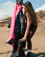Dryrobe wasserdichter Packsack in Pink und Schwarz, von Person am Strand gehalten, zeigt kompaktes Drybag-Design