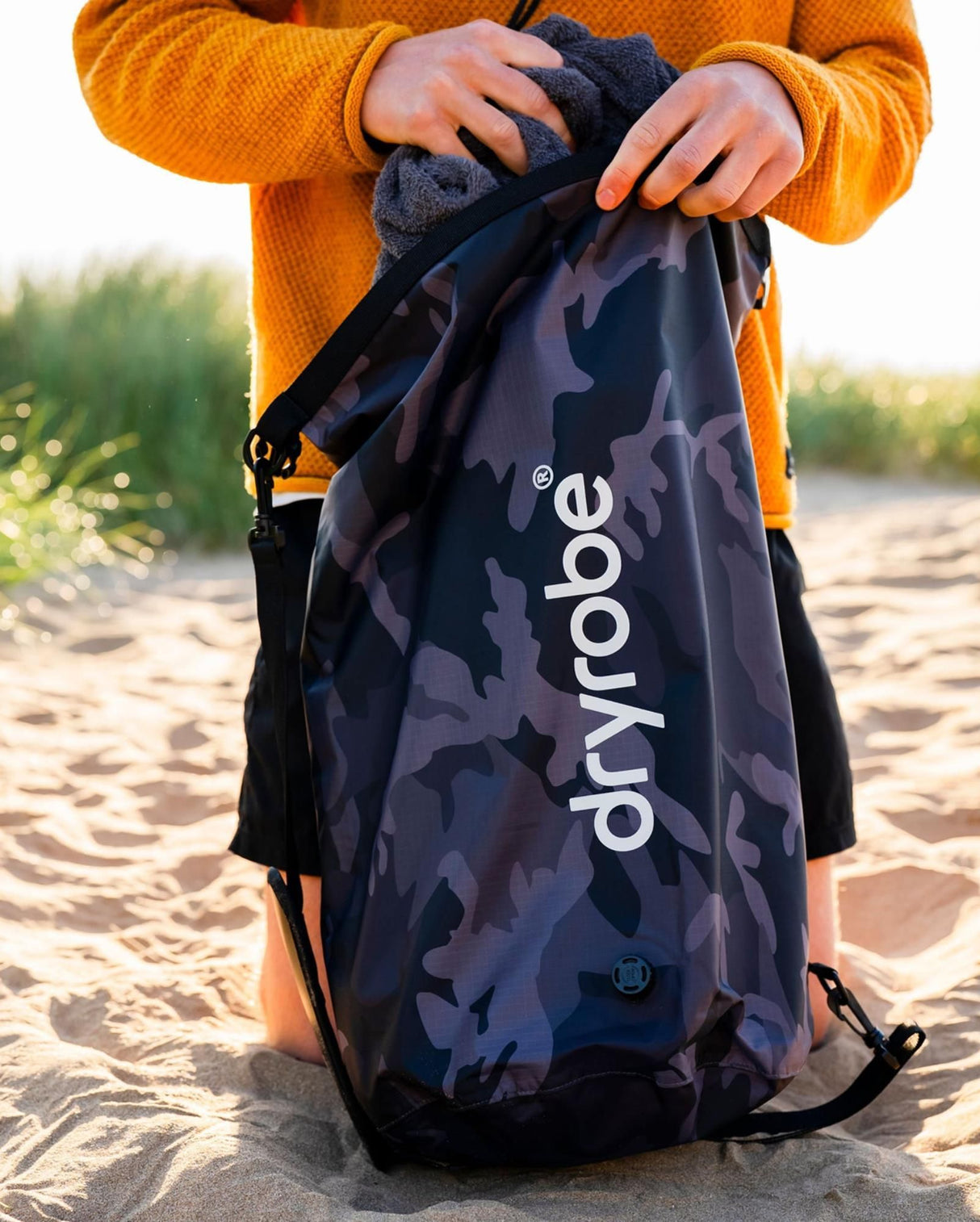 Dryrobe Drybag wasserdichter Packsack in Camouflage-Design, von Person in orangefarbener Jacke am Strand gehalten