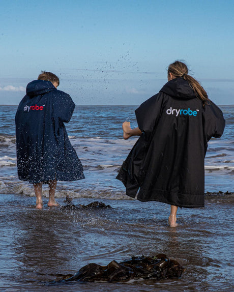 Dryrobe Advance Langarm Kinder in Marineblau und Schwarz, wasserdichte Umziehjacke aus Neopren, zwei Kinder am Strand im Meer