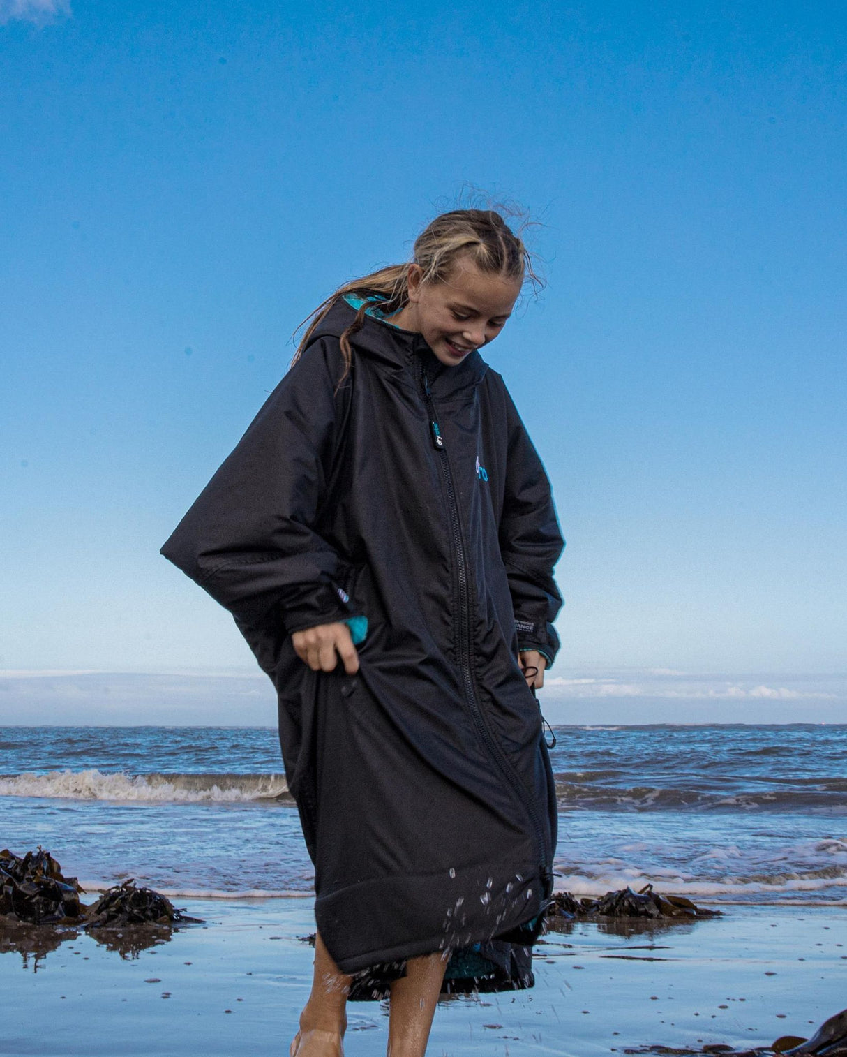 Dryrobe Advance Long Sleeve Kids schwarze Umziehtunika mit blauen Details, am Strand stehend, Frontalansicht