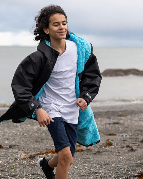 Dryrobe Advance Long Sleeve Kids schwarze Wechseljacke mit türkisem Handtuch, Junge am Strand