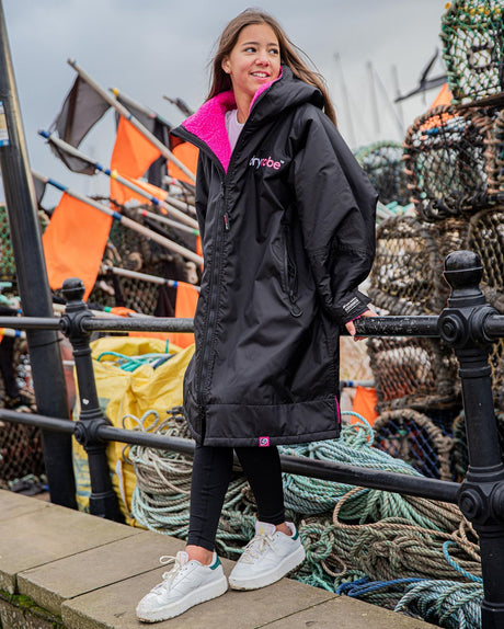 Dryrobe Advance Long Sleeve Kids schwarz mit pinkem Innenfutter, Kapuzenjacke, Vollansicht an Kind getragen am Hafen