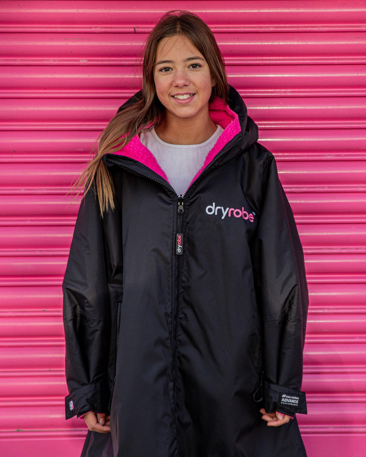 Dryrobe Advance Long Sleeve Kids schwarze Jacke mit pink gefüttertem Kapuzenausschnitt, Vorderansicht auf Kind vor pinkem Hintergrund