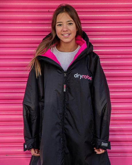Dryrobe Advance Long Sleeve Kids schwarze Jacke mit pink gefüttertem Kapuzenausschnitt, Vorderansicht auf Kind vor pinkem Hintergrund