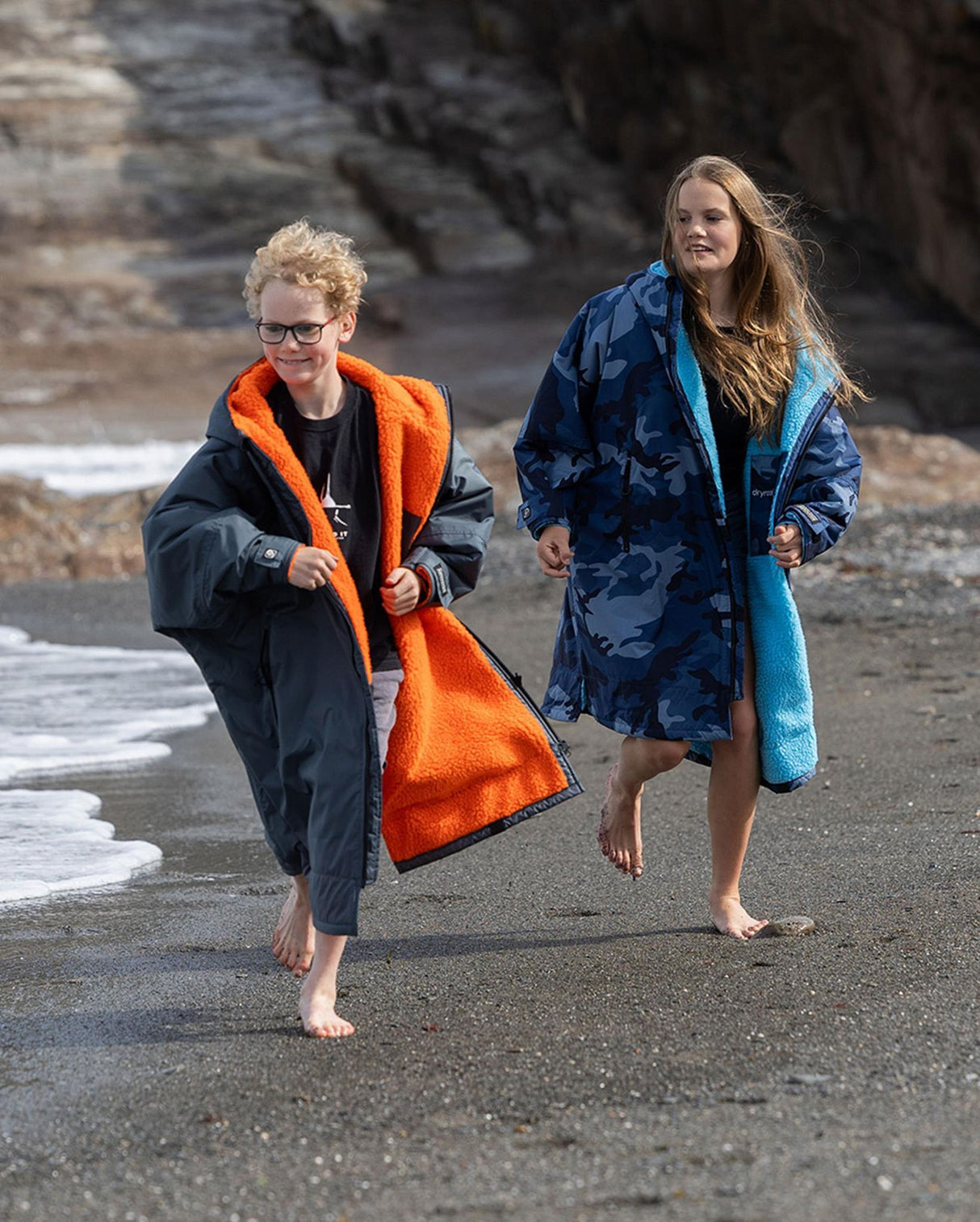 Dryrobe Advance Langarm Kids in Marineblau und Schwarz mit orangefarbenem Fleece-Futter, zwei Kinder am Strand tragend