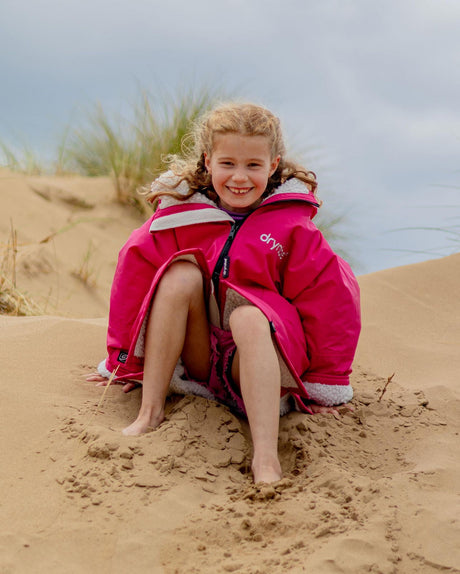 Dryrobe Advance Long Sleeve Kids in Pink mit weißen Streifen, Mädchen sitzt am Strand