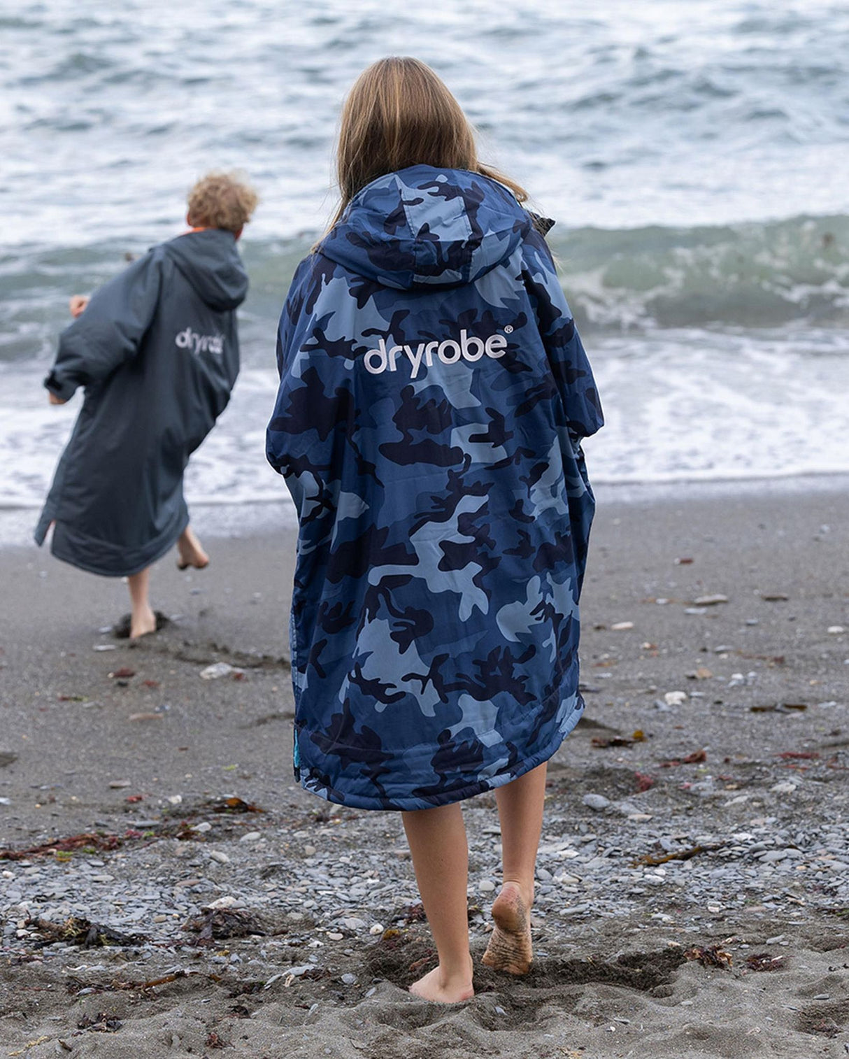 Dryrobe Advance Long Sleeve Kids in Dunkelblau mit Camouflage-Muster, getragen am Strand von hinten, Kapuze und Logo sichtbar