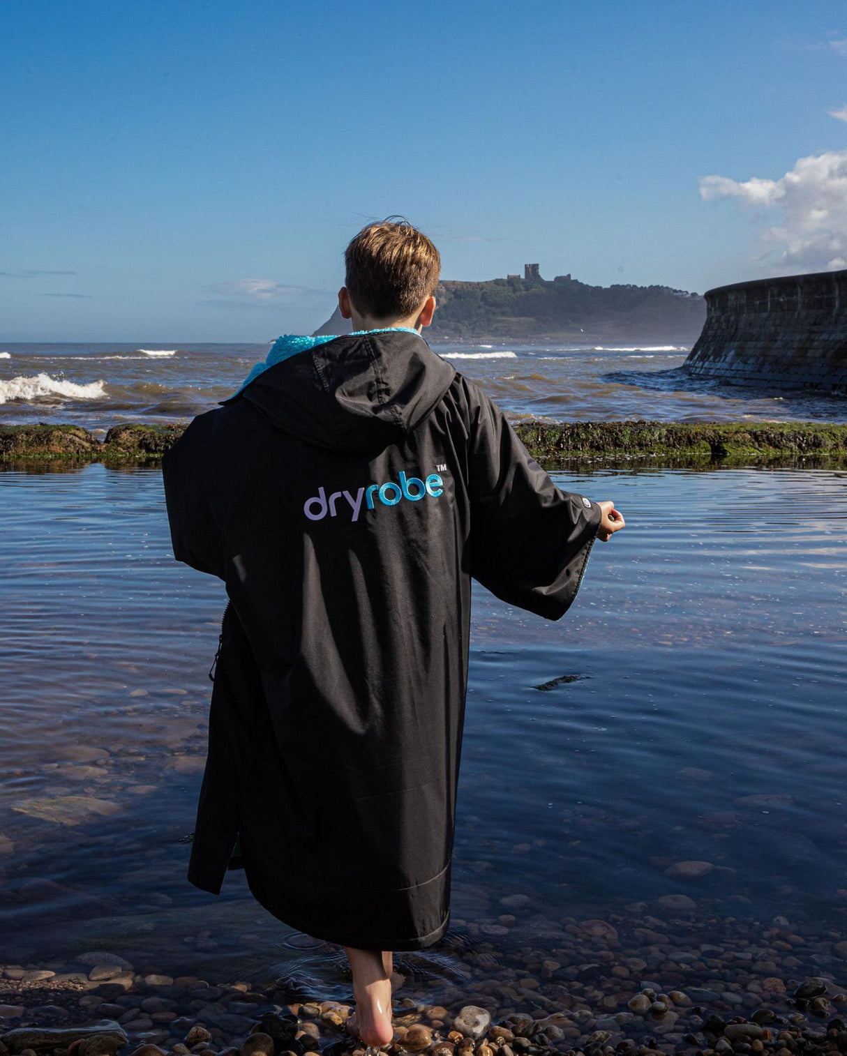 Dryrobe Advance Kurzarm Kids in Schwarz, Rückenansicht am Strand getragen, mit blauem Logo-Print
