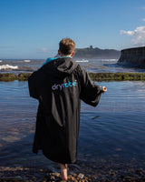 Dryrobe Advance Kurzarm Kids in Schwarz, Rückenansicht am Strand getragen, mit blauem Logo-Print