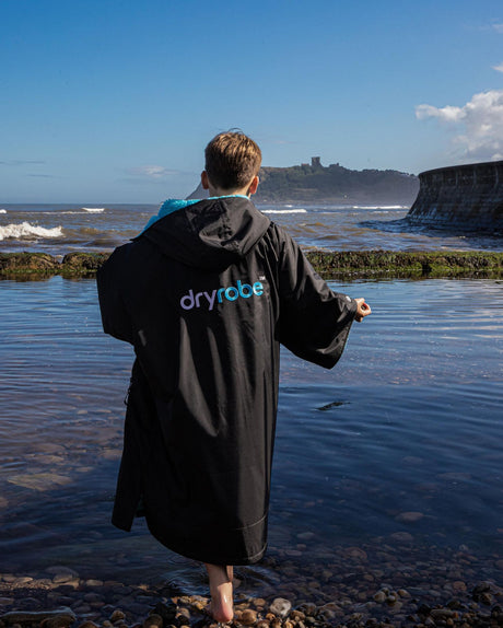 Dryrobe Advance Kurzarm Kids in Schwarz, Rückenansicht am Strand getragen, mit blauem Logo-Print