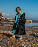 Dryrobe Advance Short Sleeve Kids Umhang in Schwarz und Türkis, Kind am Strand tragend, Frontalansicht