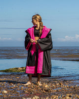 Dryrobe Advance Short Sleeve Kids in Schwarz und Pink, Mädchen am Strand trägt Bademantel mit Kapuze