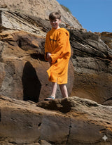 Dryrobe Kids Towel Robe Bio in Gelb, Junge trägt Bademantel aus Bio-Material an Felsen am Strand