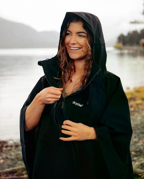 Dryrobe Regen-Poncho in Schwarz, wasserdicht, mit Kapuze, getragen von einer Frau am See