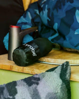 Dryrobe Mikrofaserhandtuch in Dunkelgrün, gerollt auf Holzoberfläche neben Metallbehälter, mit blauem Camouflage-Muster im Hintergrund