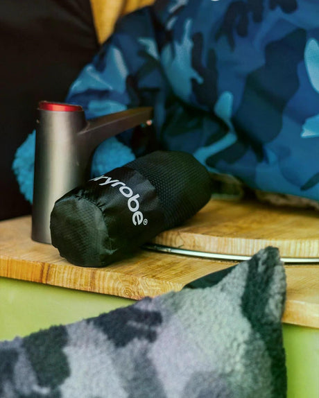 Dryrobe Mikrofaserhandtuch in Dunkelgrün, gerollt auf Holzoberfläche neben Metallbehälter, mit blauem Camouflage-Muster im Hintergrund