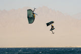Duotone Unit SLS 2027 Kiteboard grün-schwarz in Aktion über Wasser mit Kitesurfer, Frontalansicht gegen Bergkulisse