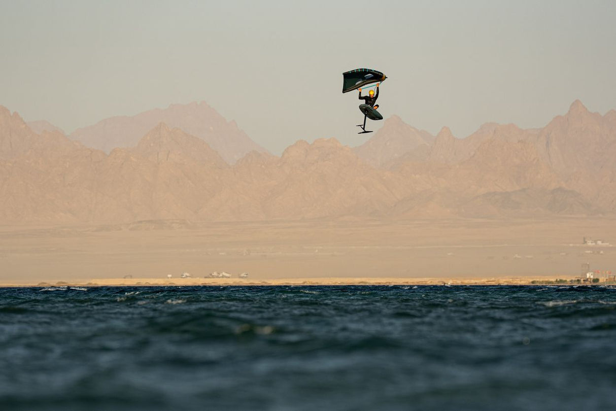 Duotone Sky Style SLS 2027 Kiteboard in Schwarz-Blau, Athlet in Aktion über Wasser mit Bergen im Hintergrund