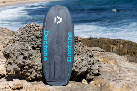 Duotone Sky Style SLS 2027 Wakeboard in Duotone-Blau, aufgestellt am Strand vor Felsen und Meer, Frontansicht