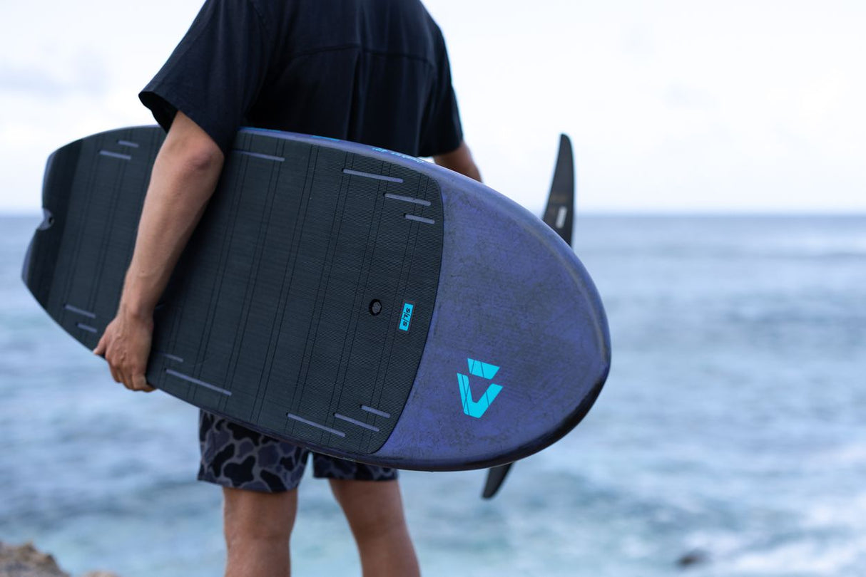 Duotone Crush SLS 2027 Kiteboard in Dunkelblau und Schwarz, von Person am Strand gehalten, Meeresansicht
