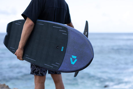 Duotone Crush SLS 2027 Kiteboard in Dunkelblau und Schwarz, von Person am Strand gehalten, Meeresansicht