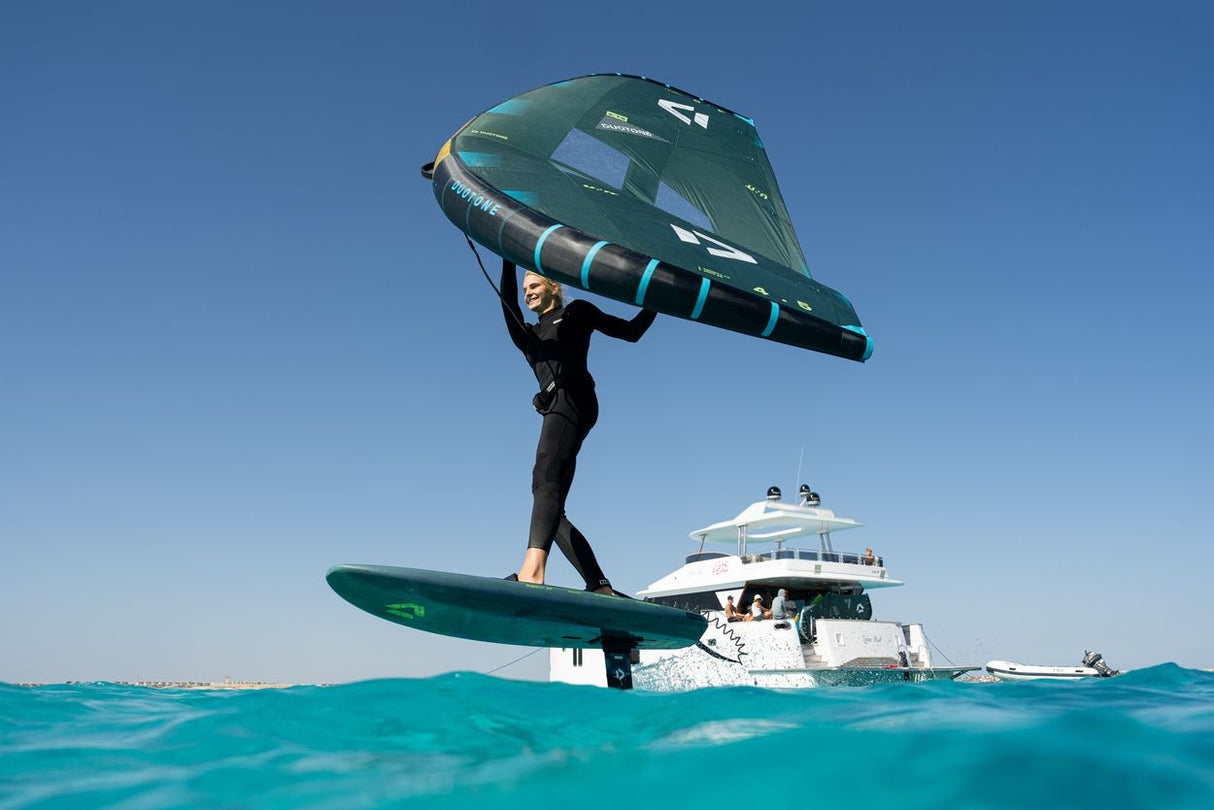 Duotone Skybrid SLS 2027 Kite in Grün-Schwarz, Kitesurfer trägt das Kite-Board über dem Wasser, Meer und Yacht im Hintergrund