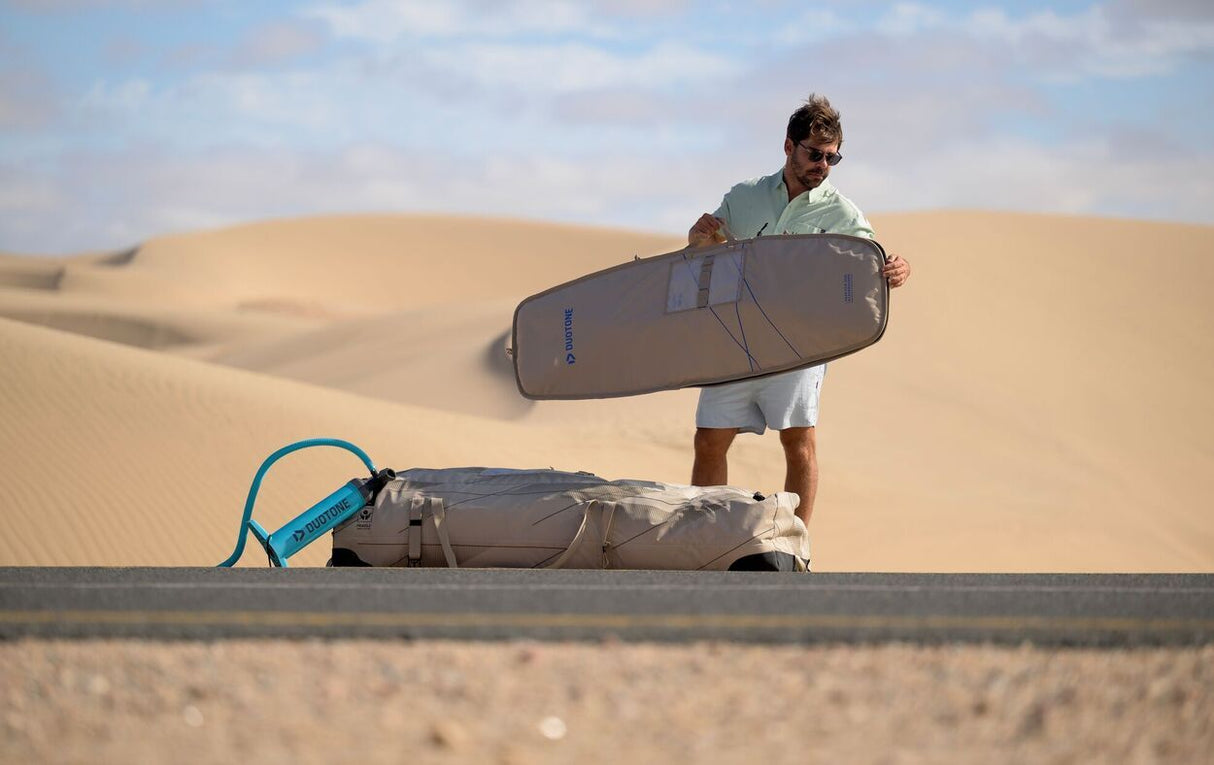 Duotone Boardbag Single Foil 2027 in Grau und Blau, Mann hält Kiteboard in Wüstenlandschaft, Tragetasche am Boden