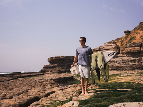 ION Graphic Tee 2026 grau, Baumwolle, Herren trägt T-Shirt am Strand mit Surfboard, Frontalansicht