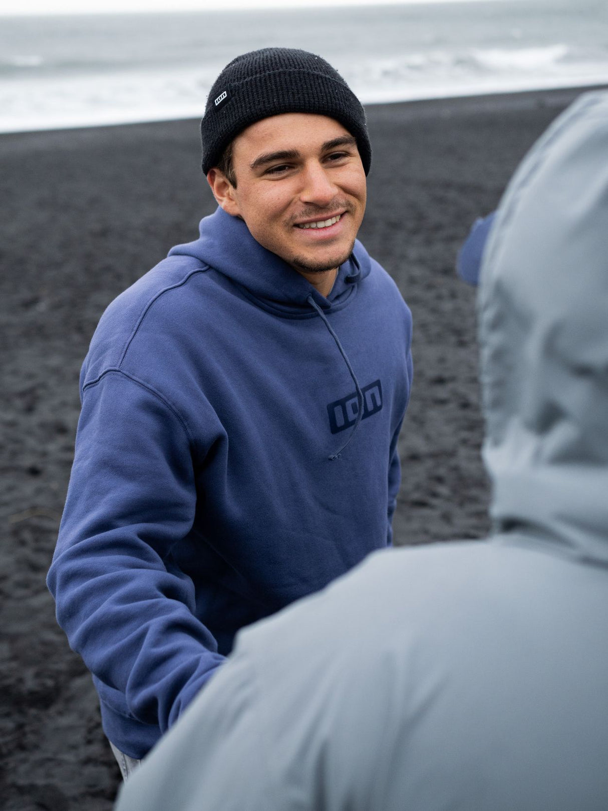 ION IONIC Hoodie 2026 in Blau, Frontalansicht auf Person am Strand mit schwarzer Mütze