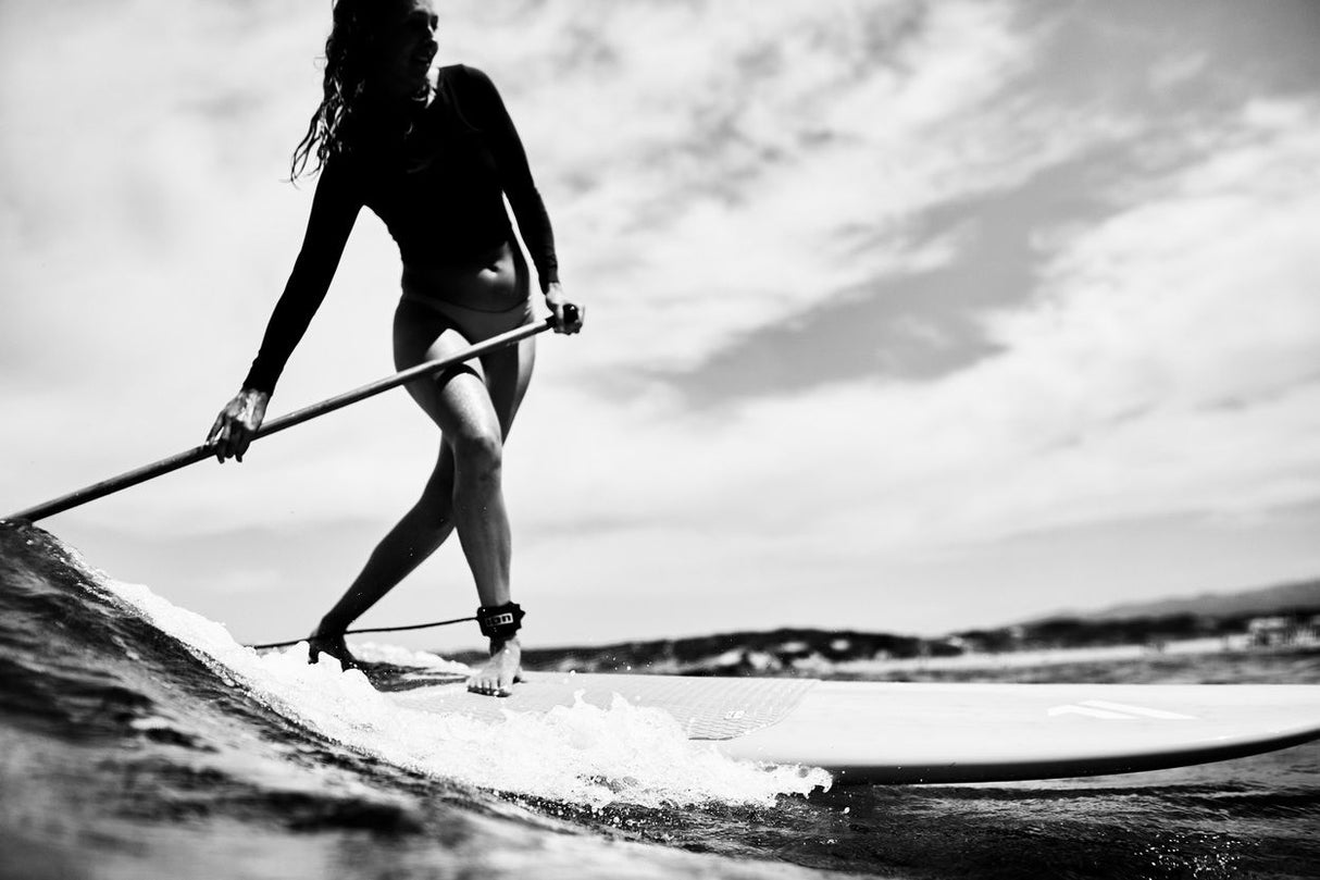 ION SUP Leash Core Ankle 2026 schwarze Fußfessel am Knöchel einer Paddlerin auf dem Wasser, Seitenansicht