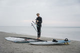 ION Harness Line 2026 schwarzes Trapez, Athlet steht mit Kiteboard am Strand, Frontalansicht, Wassersportausrüstung