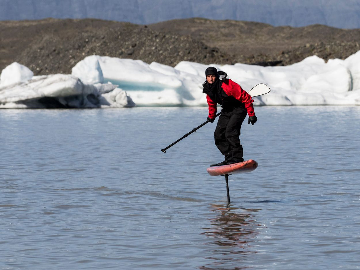 ION Fuse Lightweight Drysuit Back Zip 2026 schwarz-rot, Foiling-Action auf Wasser mit Eisbergen im Hintergrund