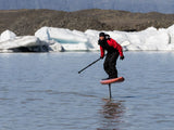 ION Fuse Lightweight Drysuit Back Zip 2026 schwarz-rot, Foiling-Action auf Wasser mit Eisbergen im Hintergrund