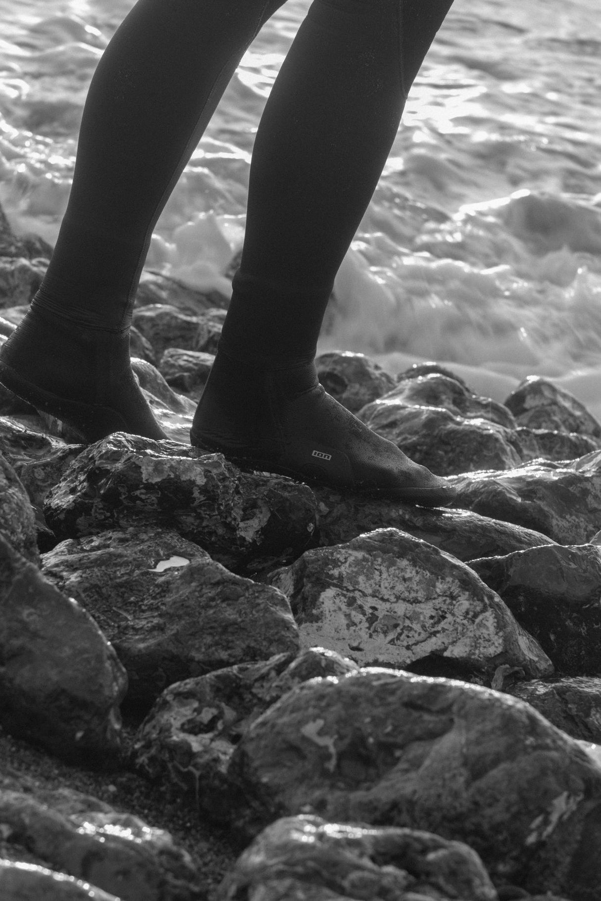 ION Ballistic Socks 3/2 Round Toe schwarze Neopren-Socken an den Füßen auf Steinen am Strand getragen