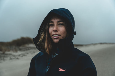 ION Neo Cosy Coat 2026 dunkelblau mit Kapuze, Frau trägt Neoprenjacke am Strand