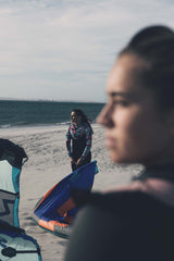 ION Ivy Weste Front Zip 2026 in Dunkelblau, Frontalansicht am Strand mit Kitesurfausrüstung