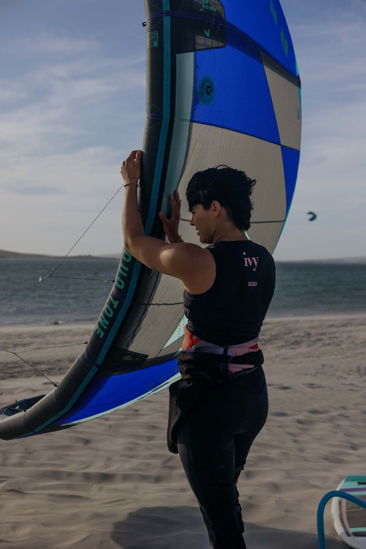 ION Ivy Weste Front Zip 2026 schwarz-blau, Neopren-Weste mit Frontverschluss, getragen beim Kitesurfen am Strand