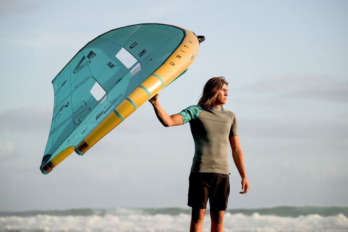 ION Neo Top 2024 Kiteboard in Türkis und Gelb, von Person am Strand getragen, Frontalansicht