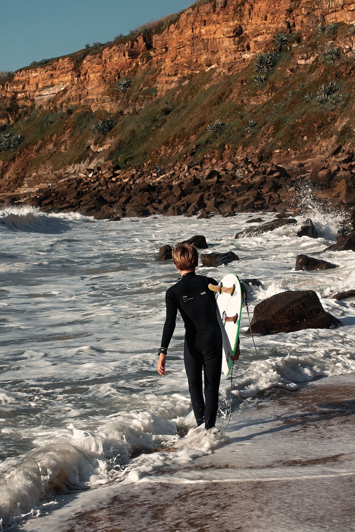 ION Capture Amp 4/3 Front Zip Junior Neoprenanzug schwarz, Kind trägt Wetsuit mit Surfboard am Strand
