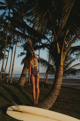ION Amaze V-Back 1.5 Damen Neoprenanzug schwarz, Frontalansicht, Frau mit Surfboard am Strand unter Palmen