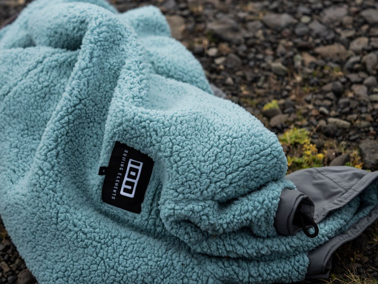 ION Storm Coat unisex 2026 in Hellblau, Fleece-Material, Detail-Ansicht mit ION-Logo auf steinigem Untergrund