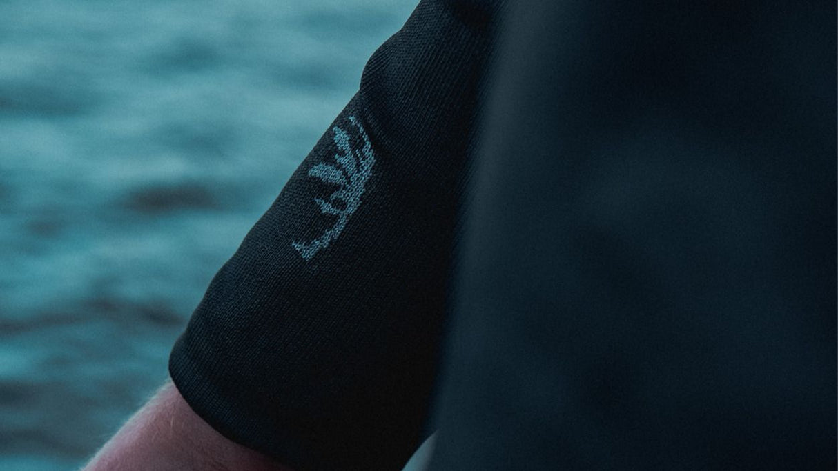 ION Rashguard LS Surf Shield SS2026 schwarzer Langarm-Neoprenanzug mit Logo-Detail am Arm, vor Wasser-Hintergrund