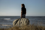 ION Wing Boardbag Core 2026 Transporttasche in Beige, Mann trägt Boardbag am Strand mit Meer im Hintergrund