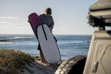 ION Prone Foil Boardbag Core 2026 in Bordeaux und Grau, geöffnet am Strand, Mann trägt Board, Frontalansicht mit Meer im Hintergrund