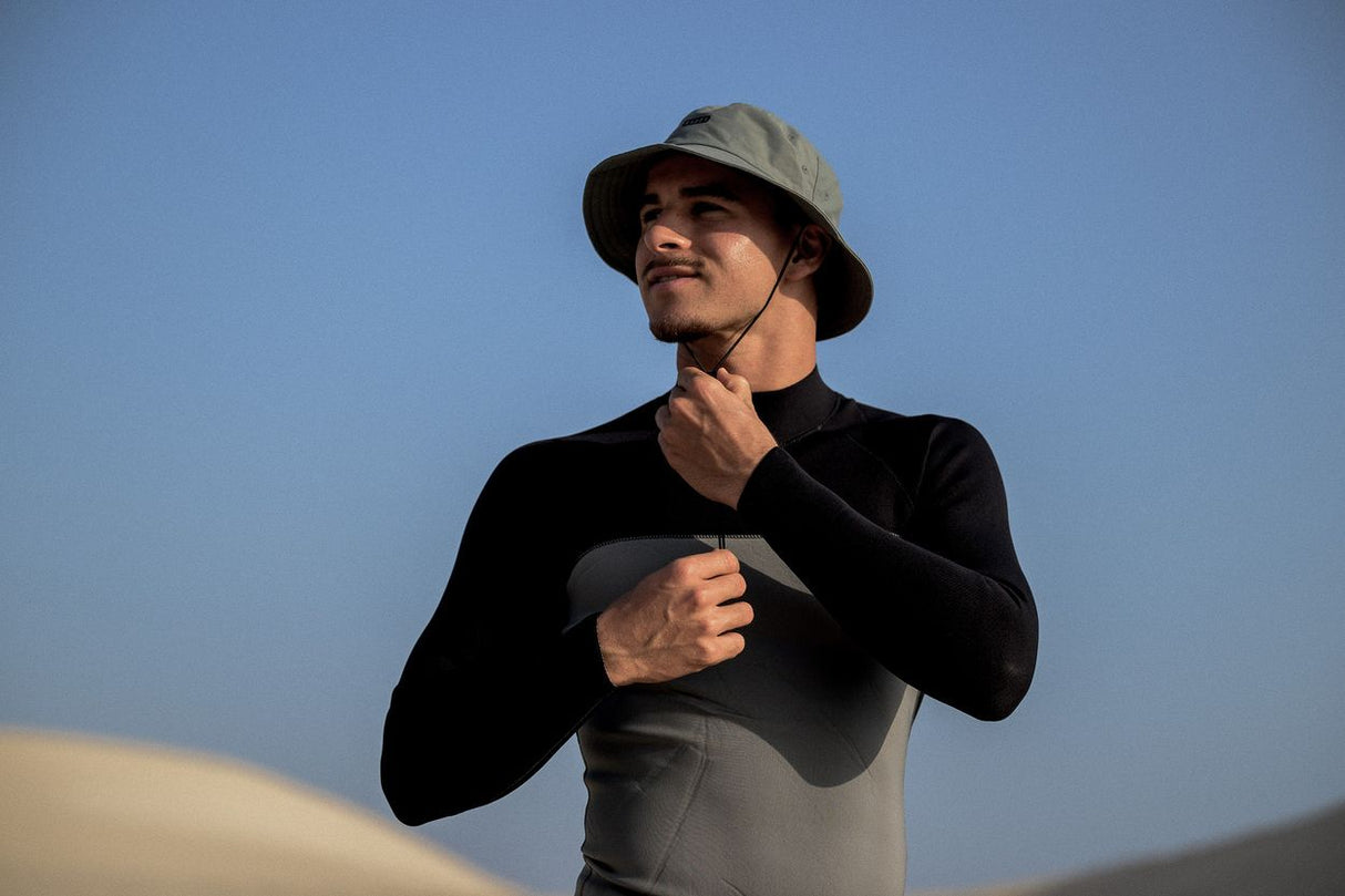 ION Bucket Surf Hat 2024 in Grau, getragen von Mann mit Kinnriemen, Frontalansicht vor blauem Himmel