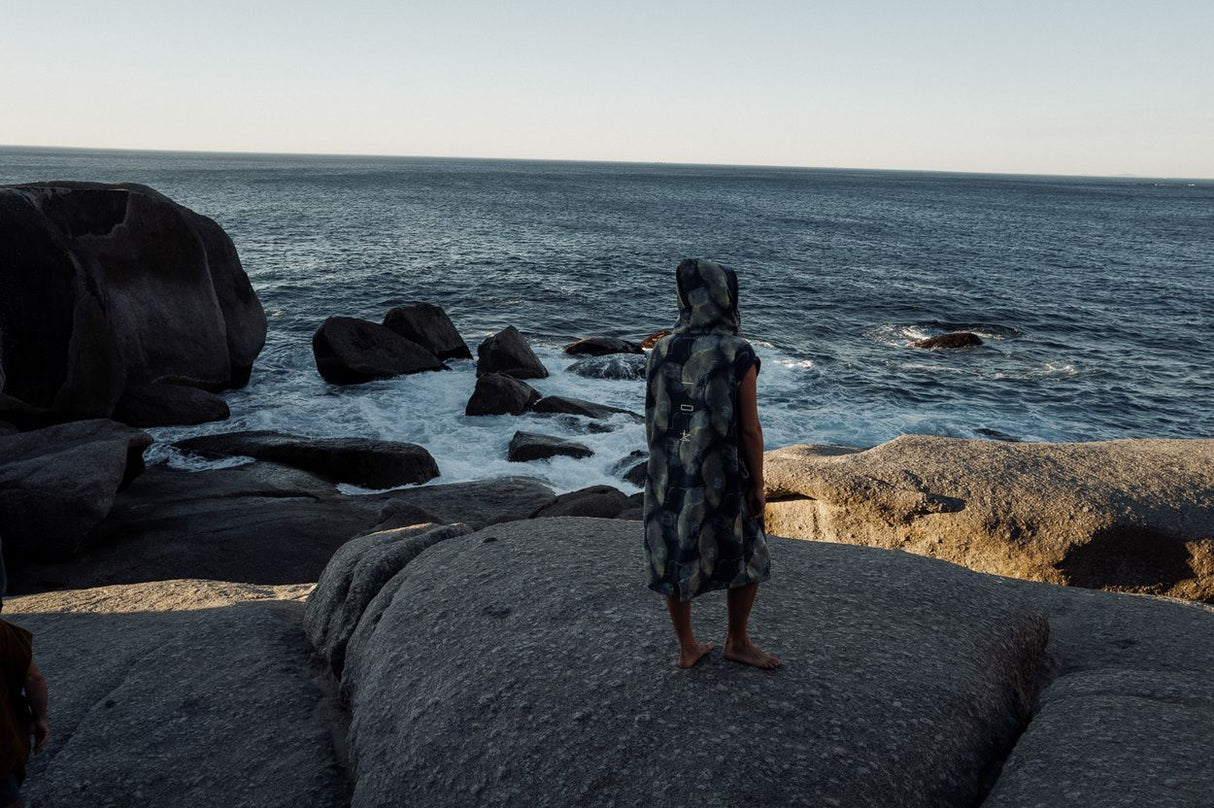 ION Surf Poncho Element 2026 schwarz, Person trägt Poncho am Strand mit Felsen und Meer
