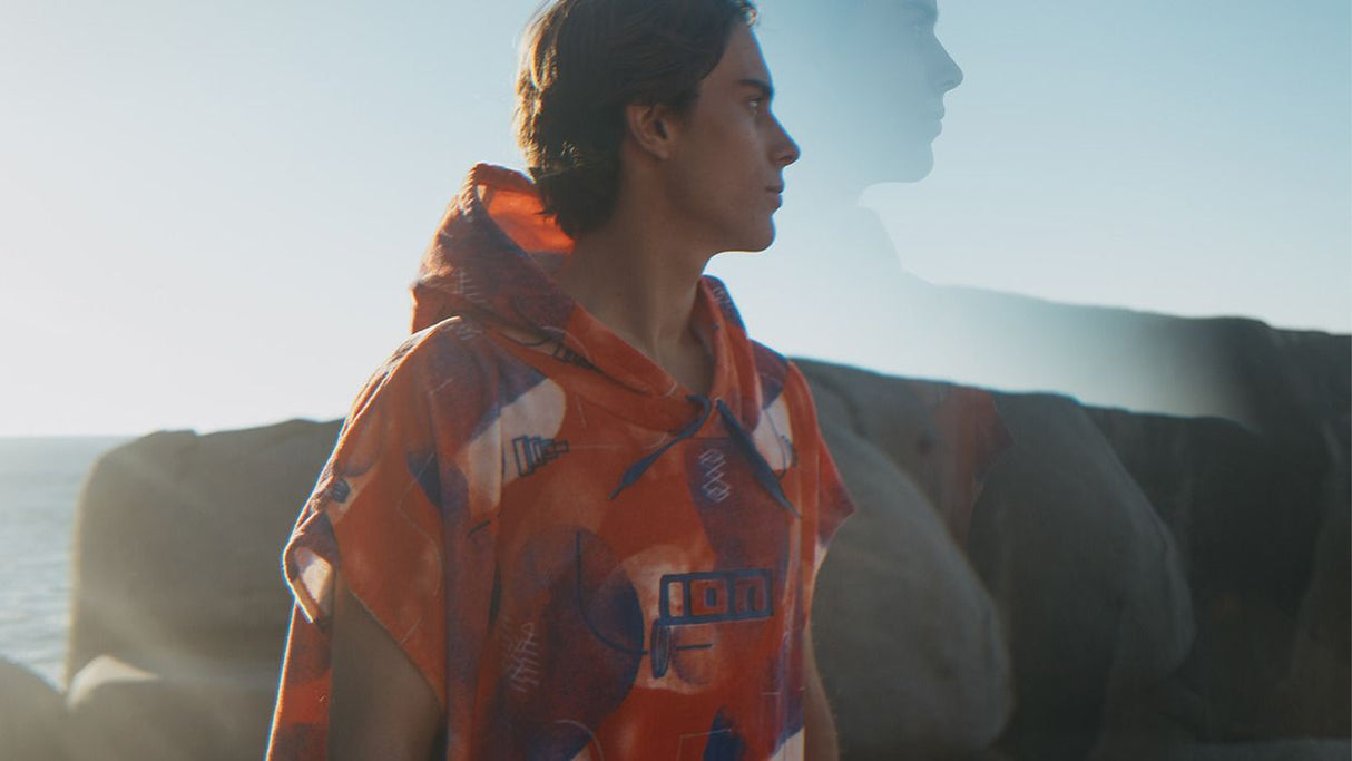 ION Poncho Core Lumen 2026 in Rot mit Kapuze, Seitenansicht einer Frau am Meer tragend