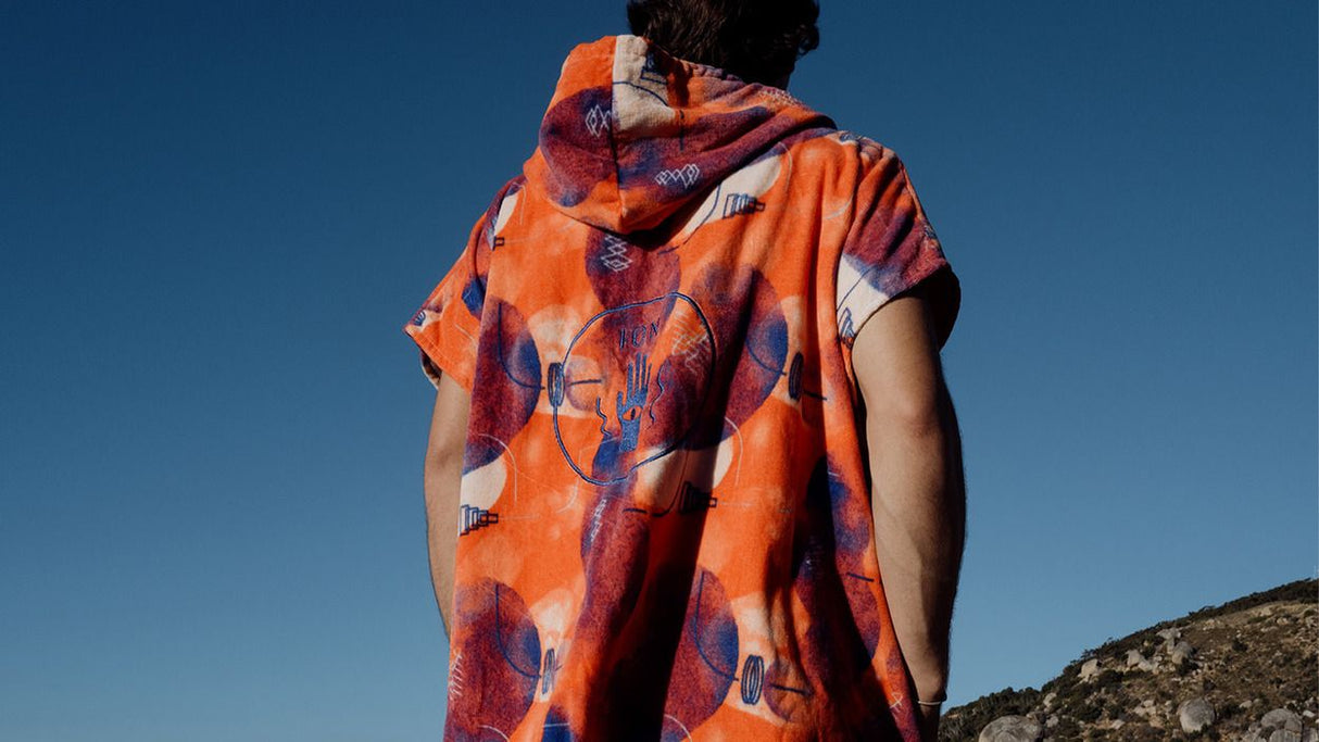 ION Poncho Core Lumen 2026 orange mit geometrischem Muster, Baumwolle, Rückansicht auf Person vor blauem Himmel