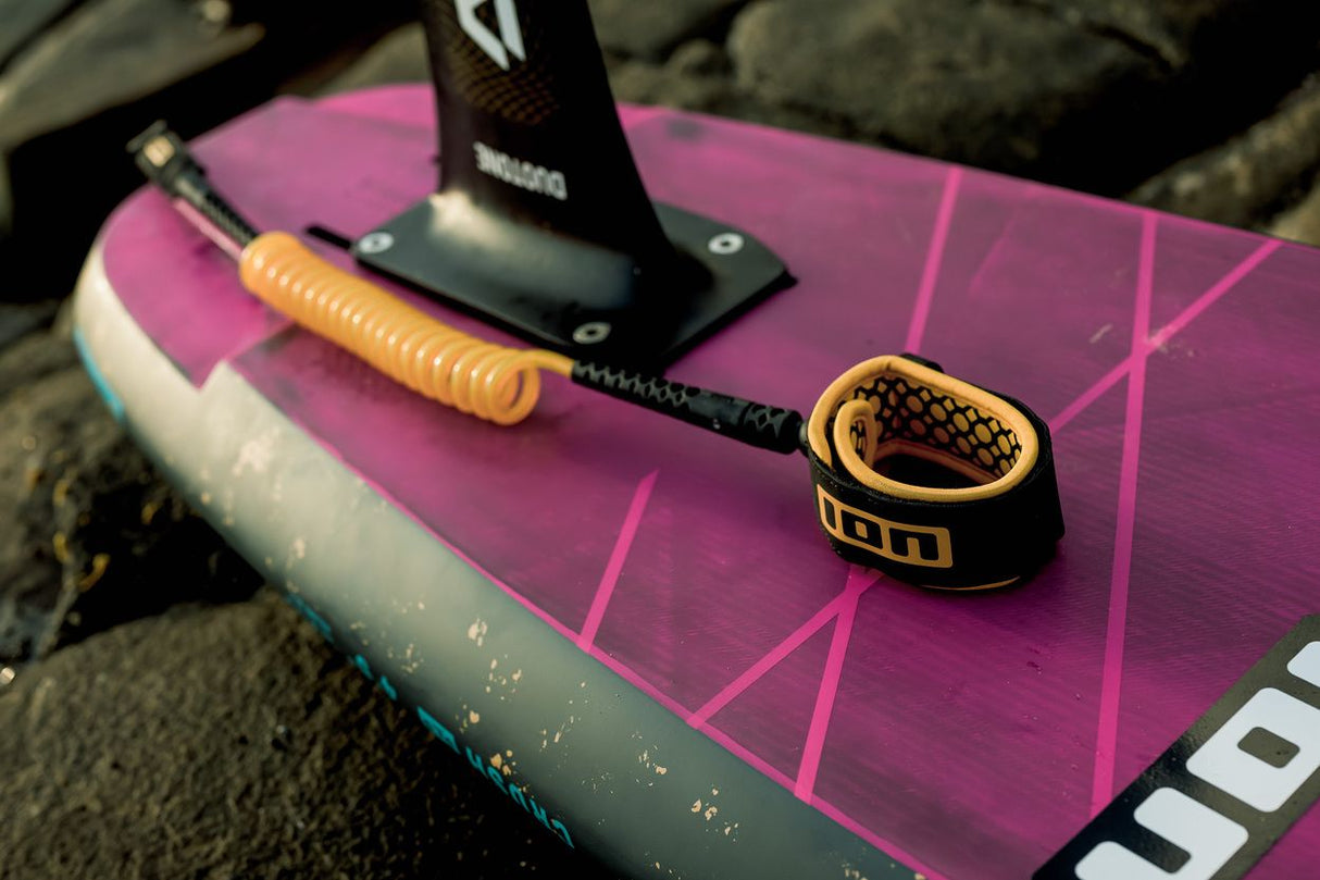 ION Wing Board Leash Core Coiled Knee schwarz-gold, aufgerollt auf pink-weißem Board, Draufsicht